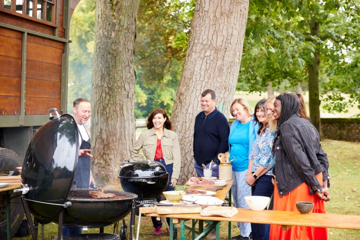 Group of people standing by a grill under trees at a park.