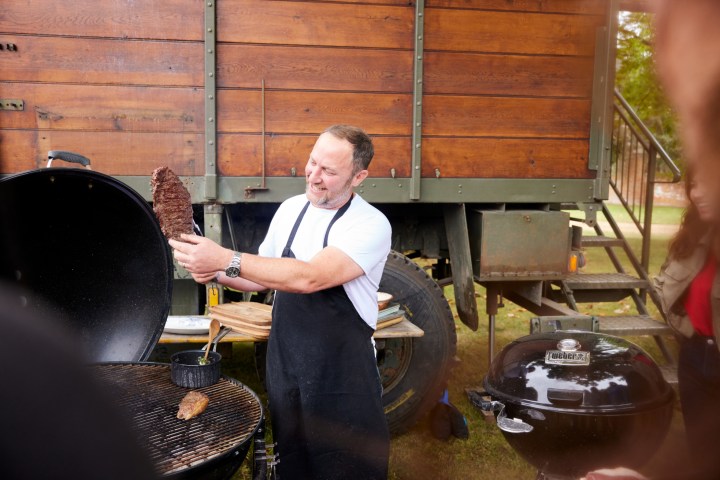 Man grilling meat near a rustic vehicle outdoors.