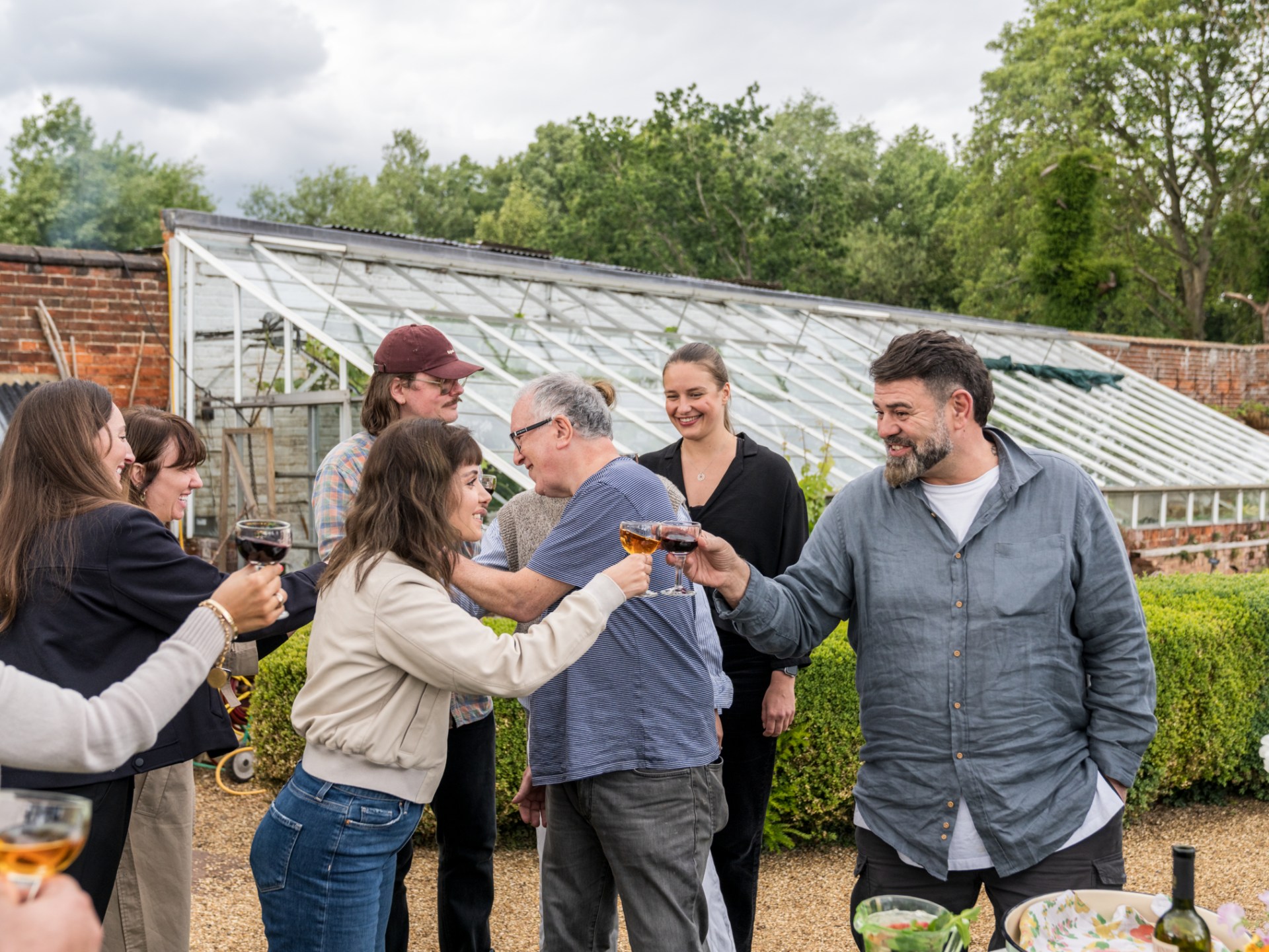 Group of people toasting with drinks outside near a greenhouse.