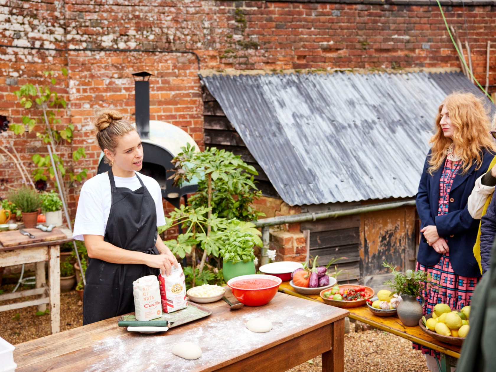 Outdoor cooking demo with a chef and three spectators in a rustic setting.