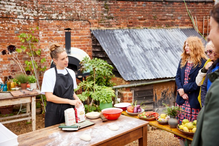 Outdoor cooking demo with a chef and three spectators in a rustic setting.