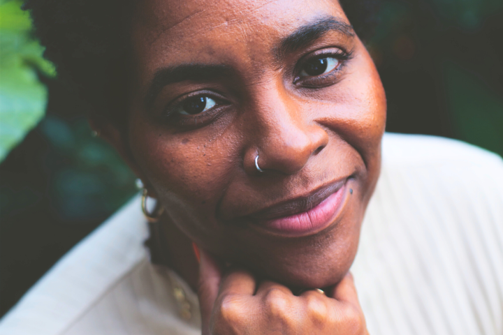 Woman with short hair and a nose ring smiling, hand on chin, wearing a light sweater.