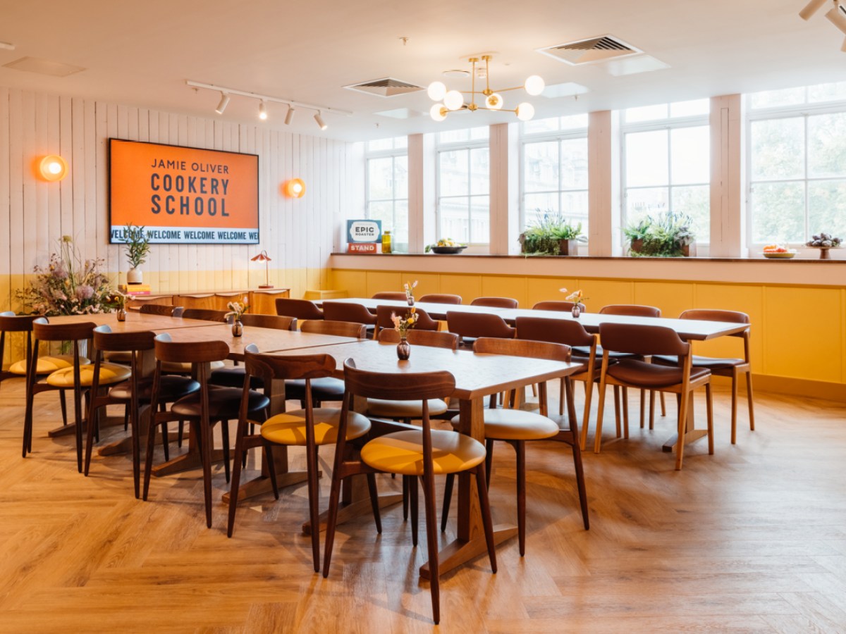 Cookery school room with long tables, chairs, plants, and a sign for Jamie Oliver Cookery School.