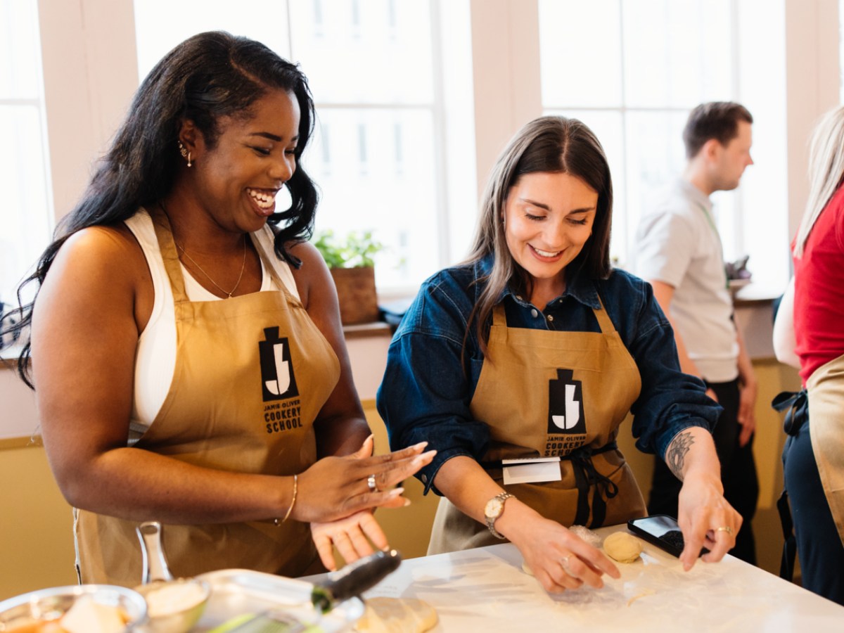 Two women smiling and shaping dough in a cooking class, wearing beige aprons.