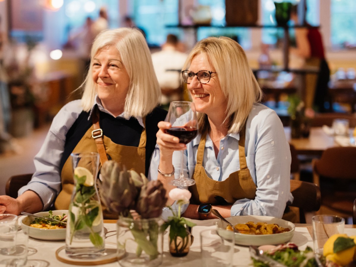 Two women in aprons smile at a restaurant table with plates of food and a glass of wine.