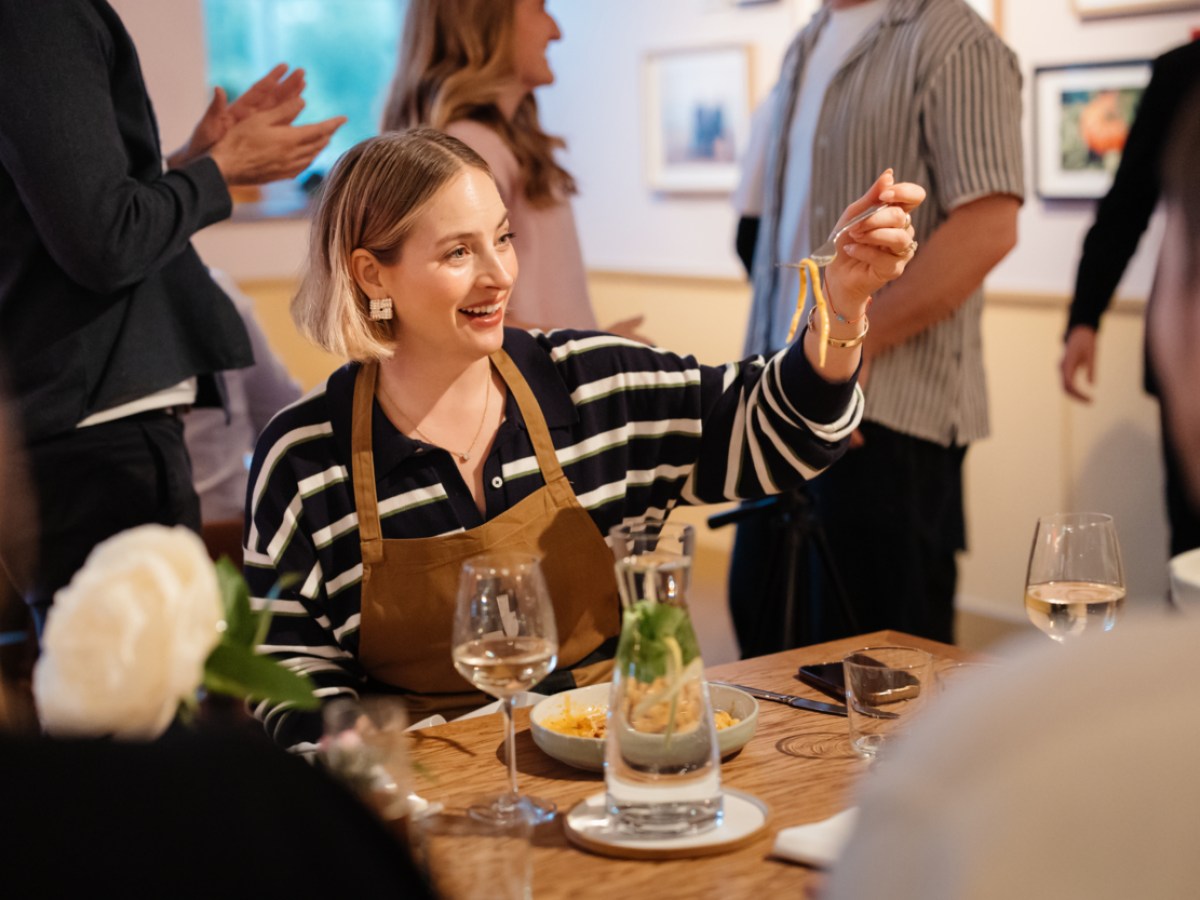 Smiling woman in striped shirt holding pasta at a dining table during a social gathering.