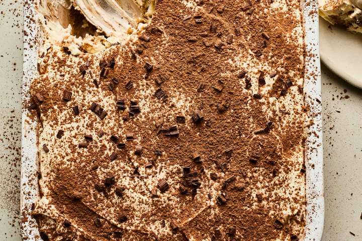 Tiramisu with cocoa on top, partially served, spoon in dish, slice on plate.