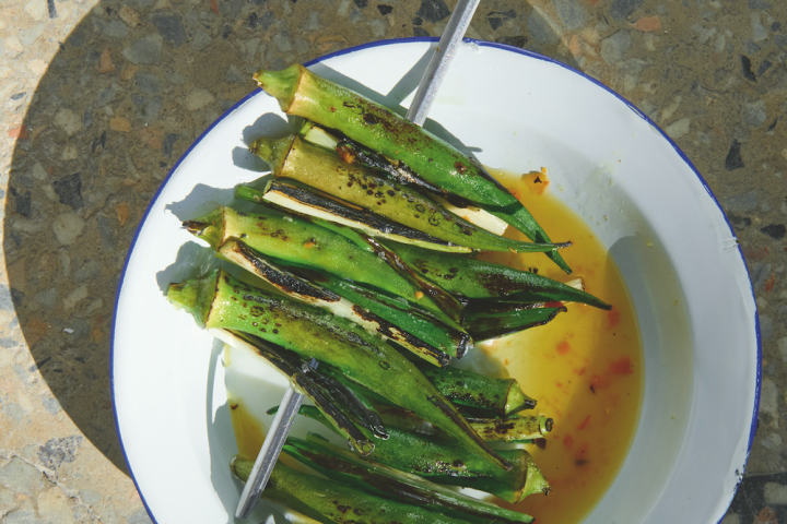 Grilled okra on skewers in a bowl with sauce on a speckled surface.
