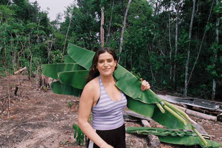 Person holding large green leaves in a forest clearing, wearing a purple tank top.
