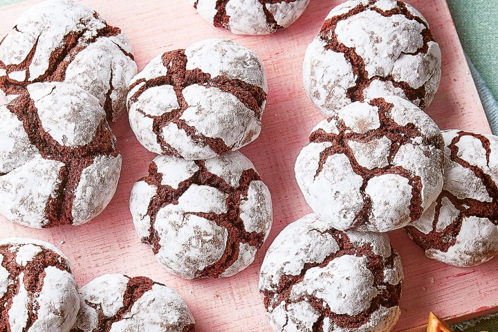 Chocolate crinkle cookies dusted with powdered sugar on a pink board, with decorated star and tree cookies nearby.