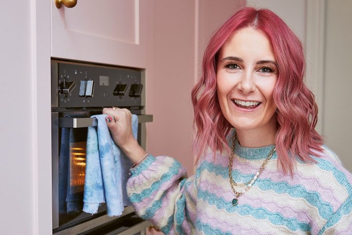 Smiling person with pink hair holding a tray of baked goods in a kitchen with pink cabinets.
