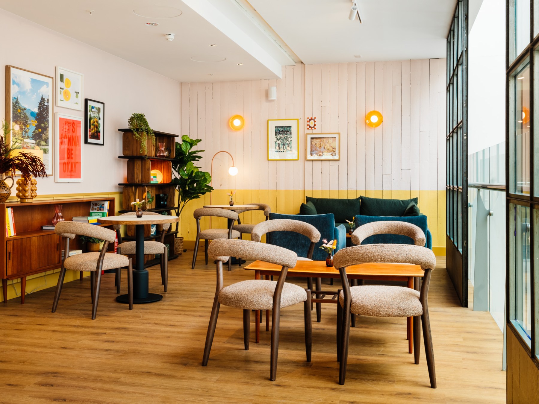 Cozy cafe interior with wooden tables, upholstered chairs, artwork on walls, and large windows.