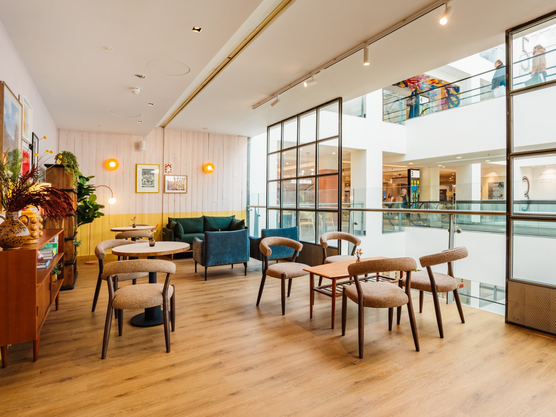 Modern lounge with wood flooring, round tables, chairs, and wall decor, adjacent to an indoor balcony.