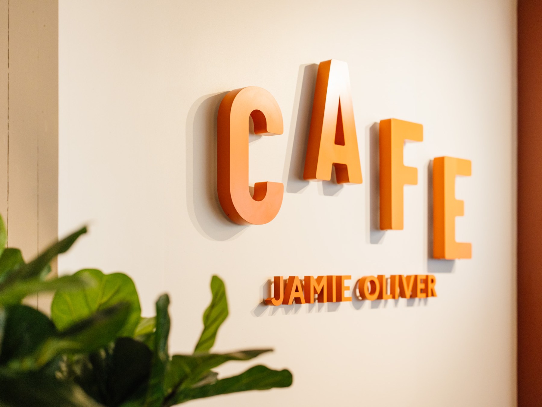 Orange 'CAFE' letters on a wall with 'JAMIE OLIVER' below, beside green plants.