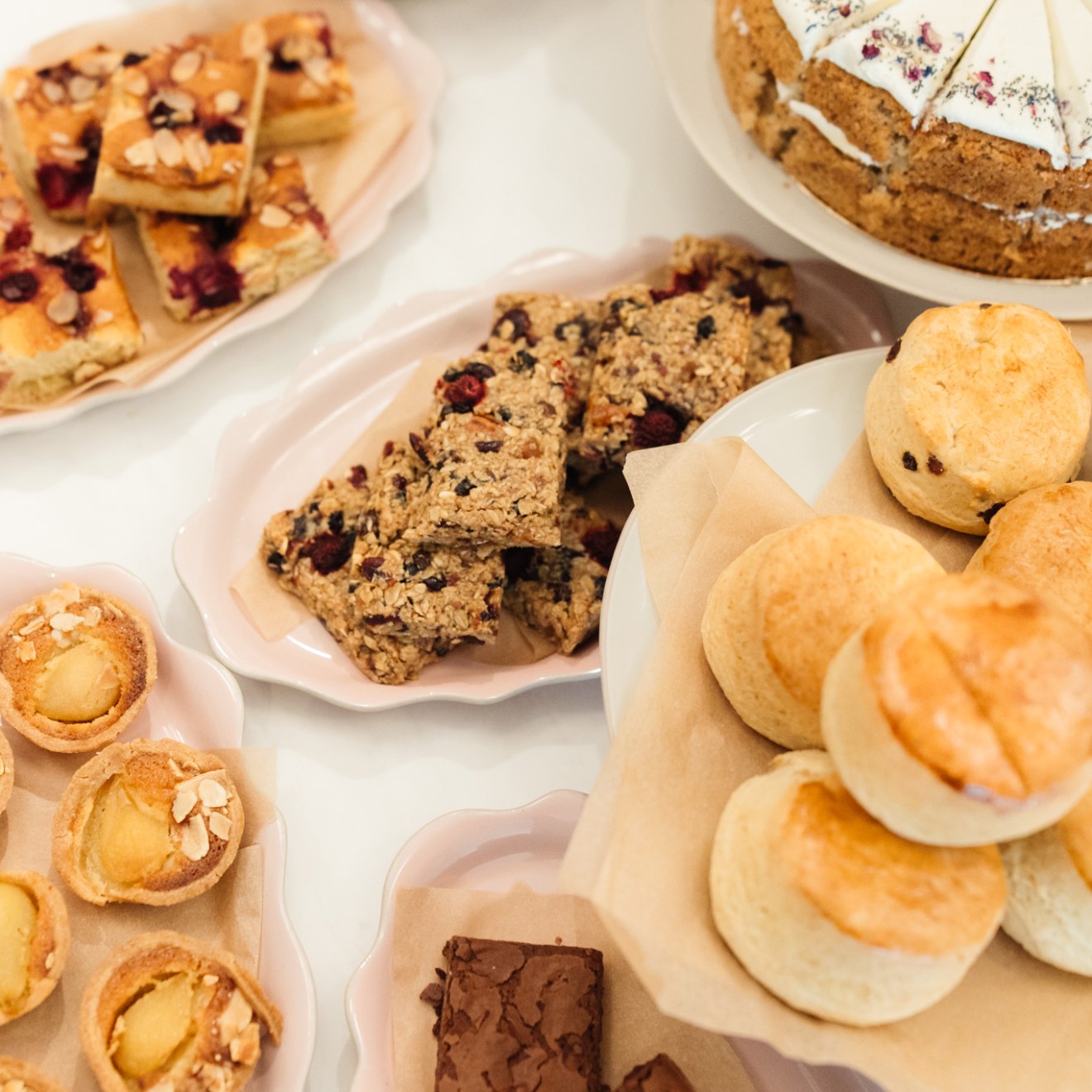 web__017__jamie-oliver-cookery-school-launch__cafe_9b6ad3 Assorted pastries and cakes on plates, including slices of cake, scones, and fruit-topped tarts.