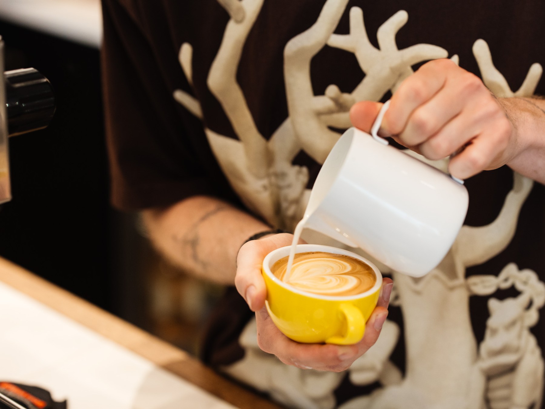 Barista pouring latte art heart into a yellow cup.