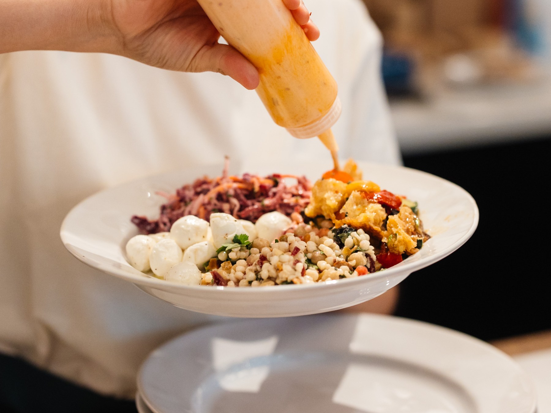 Hand pouring sauce over a plate with grains, cheese, and vegetables.
