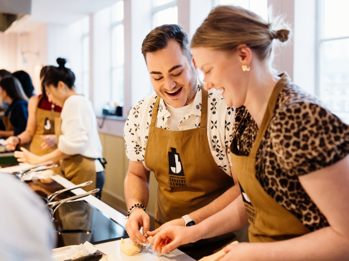 People in aprons smiling and preparing food together in a bright kitchen.