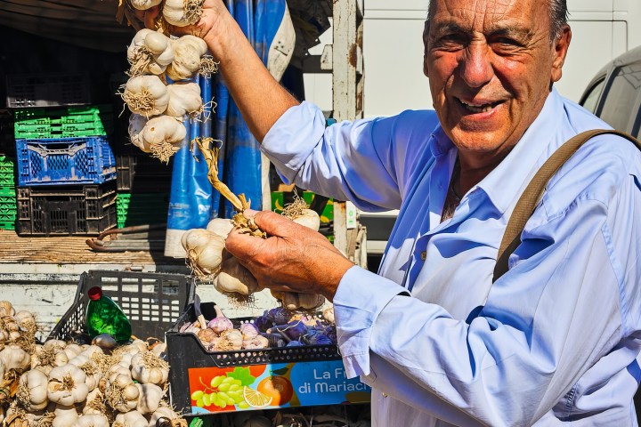 Man holding bunches of garlic at an outdoor market with crates in the background.