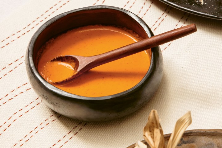 Bowl of orange soup with wooden spoon on a patterned cloth, tamales in husks on plates nearby.