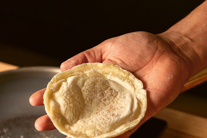Hand holding a small, uncooked pizza dough above a pan.