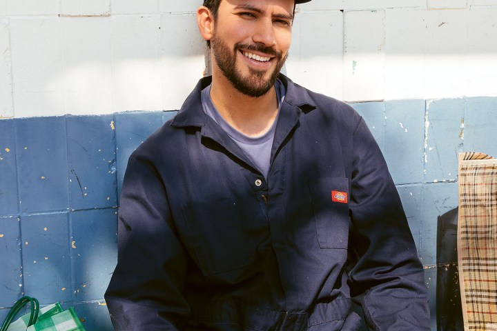 Smiling man in blue coveralls and cap sitting by bags.