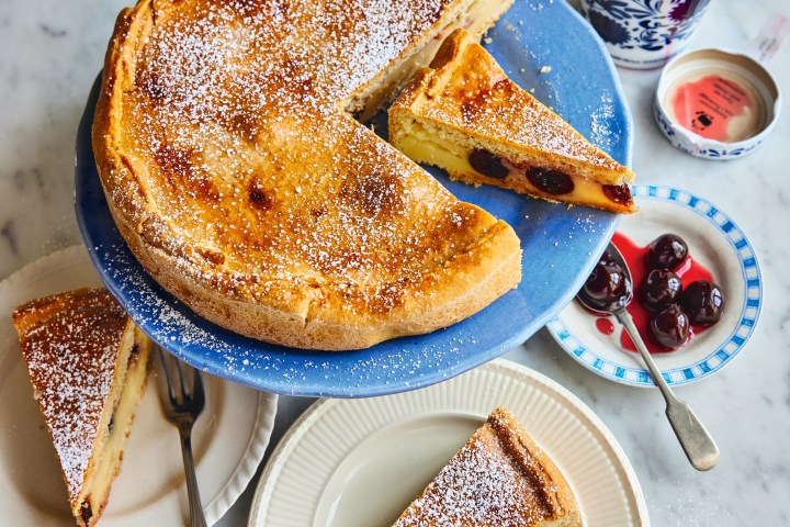 Cherry cake on a blue platter with powdered sugar, sliced with plates and a jar of cherries nearby.