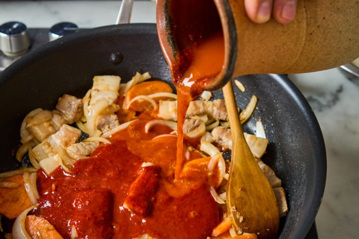 Hand pouring sauce into a skillet with meat and vegetables on a stove.