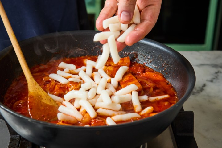 Person adding tteok to a pan of red sauce on a stove.