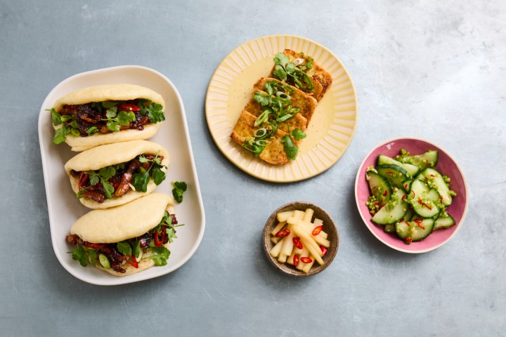 Plates of bao buns, tofu, cucumber salad, and pickled radish on a gray surface.