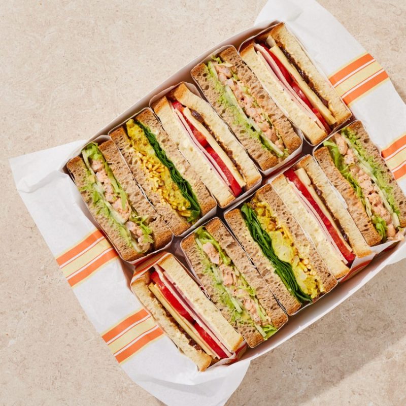 Box of assorted sandwiches cut in halves on paper.