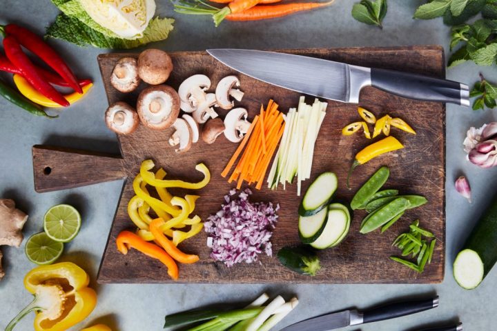 Wooden board with sliced vegetables and knives, surrounded by whole veggies.
