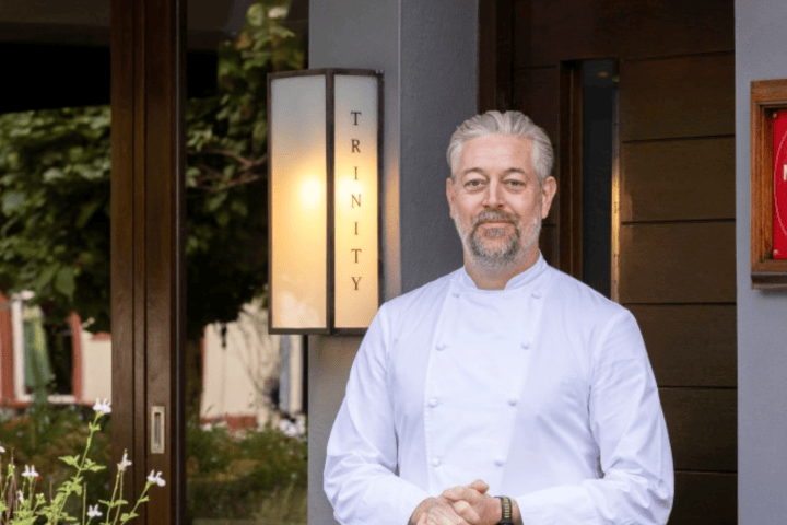 Chef in white uniform standing outside a restaurant with a 'Trinity' sign.